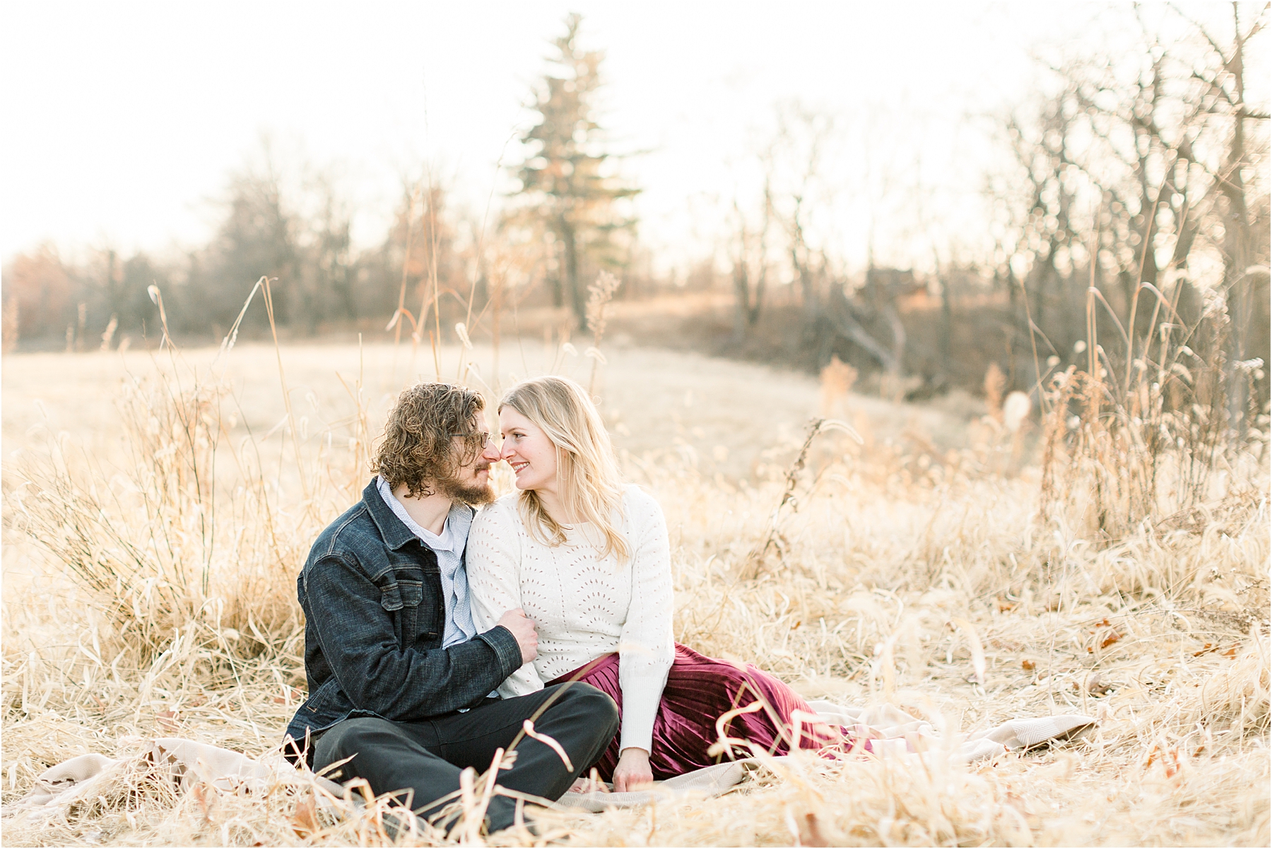man in jean jacket and girl in sweater and velvet skirt cuddling in grass for winter anniversary session